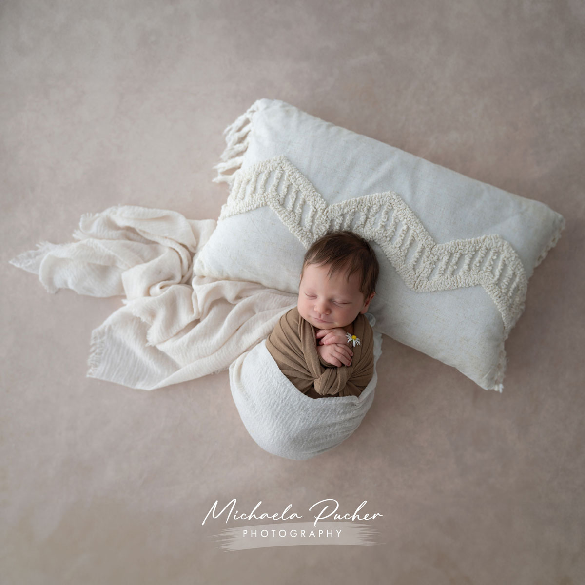 Schlafendes Neugeborenes, eingewickelt in braune und weiße Tücher, liegend auf einem großen weißen Kissen mit Boho-Struktur und umgeben von weichen Stofflagen.